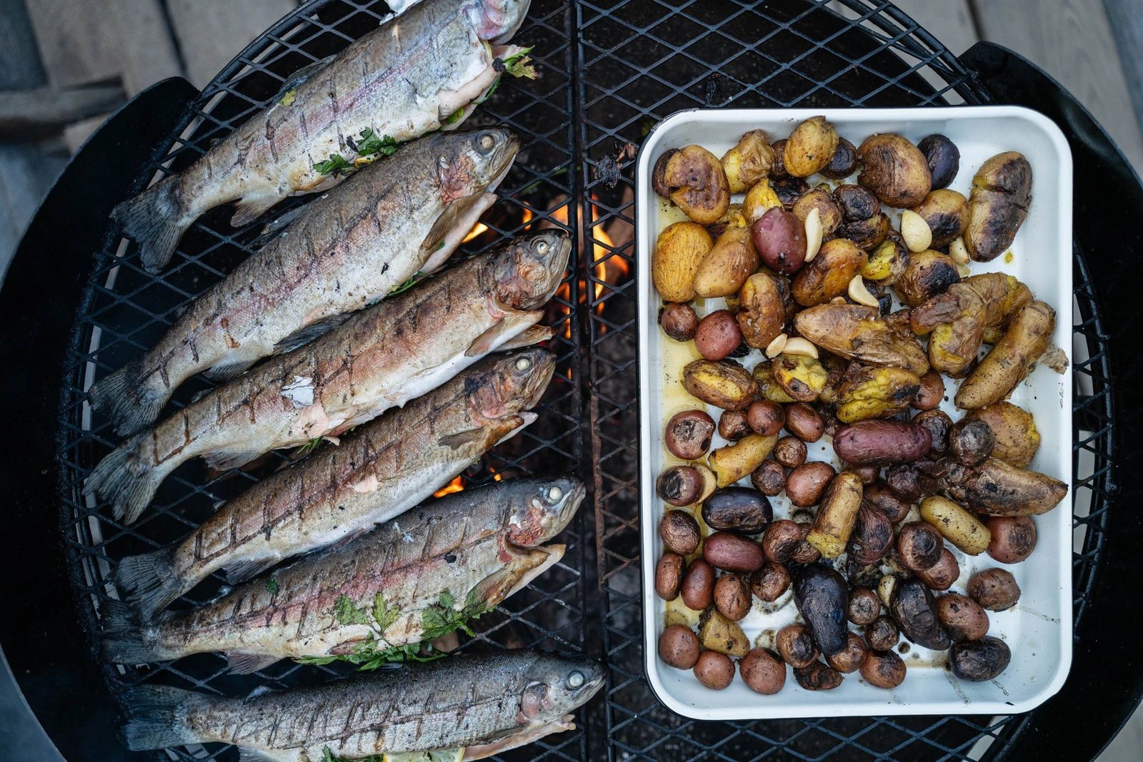 Whole trout cooking over open fire with roasted potatoes